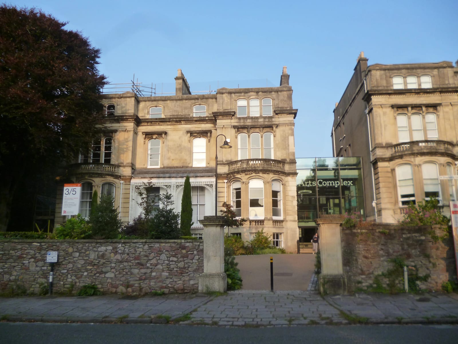 The main entrance to the Arts Complex at the University of Bristol. Two large limestone buildings are connected by a modern glass atrium, set in a courtyard with trees and bushes.