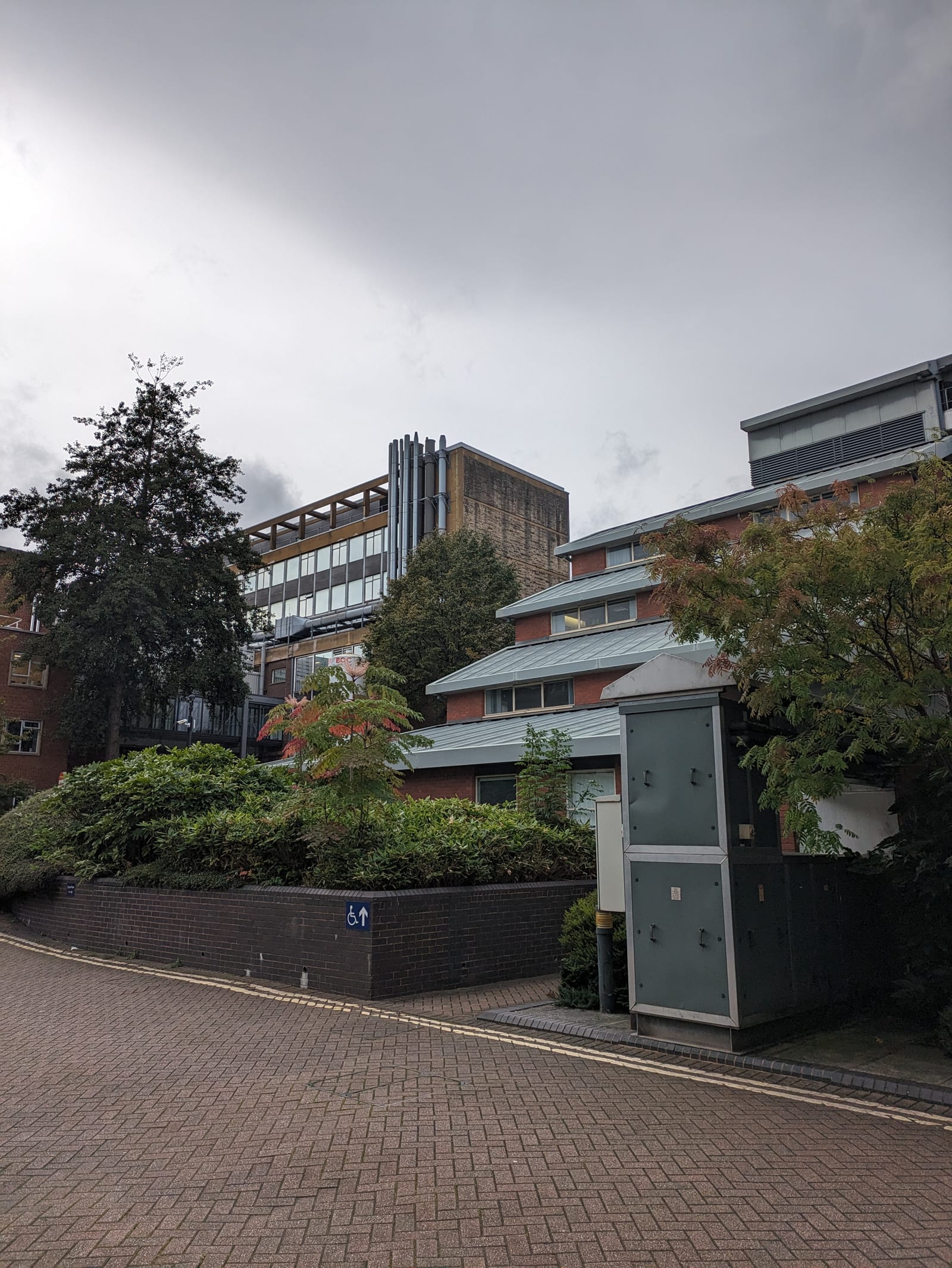 Landscape taken on an overcast afternoon. A brick paved road is in the foreground. Behind it, a building with multiple staggered floors and sloping grey rooves is partly occluded by trees, shrubs in a brick planter and a large electrical box. In the background the corner of a much taller building is visible with many metal chimneys grouped together on one edge.